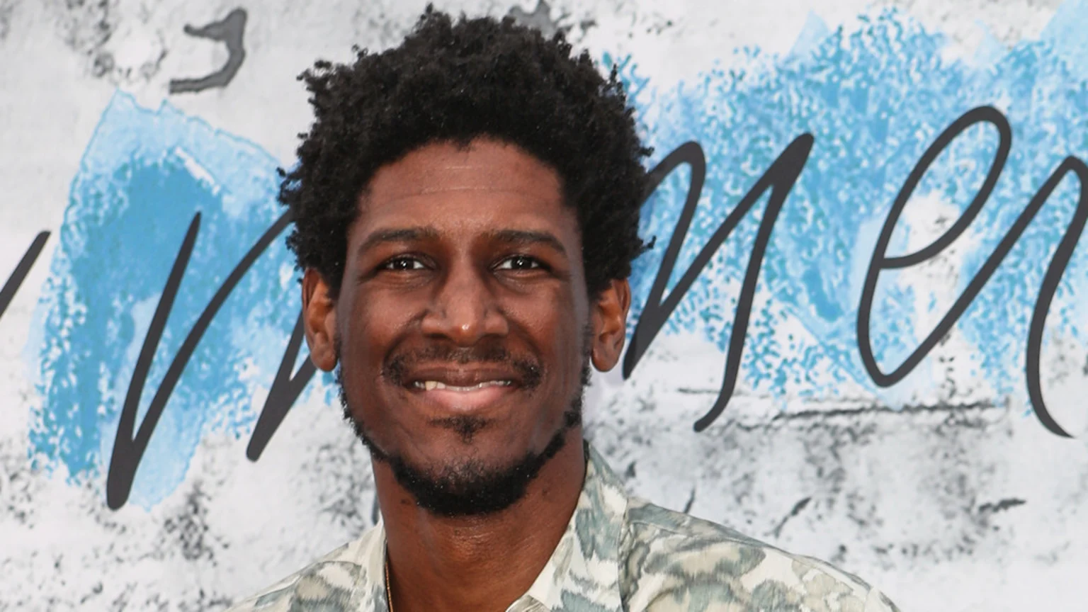 A smiling man with dark curly hair and a short beard wears a patterned shirt, standing against a white background with blue and black abstract art and cursive writing.