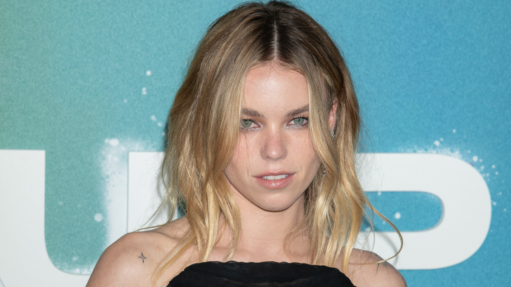Milly Alcock, a young woman with wavy blonde hair, green eyes, and freckles, looks at the camera, wearing a dark top, with a star tattoo on her left shoulder against a blue background.