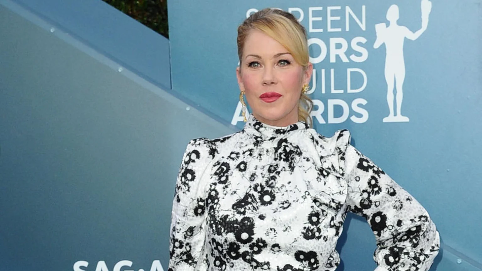 Christina Applegate poses at an event, wearing a black and white floral top, gold earrings, and red lipstick, against a blue backdrop with the SAG Awards logo.