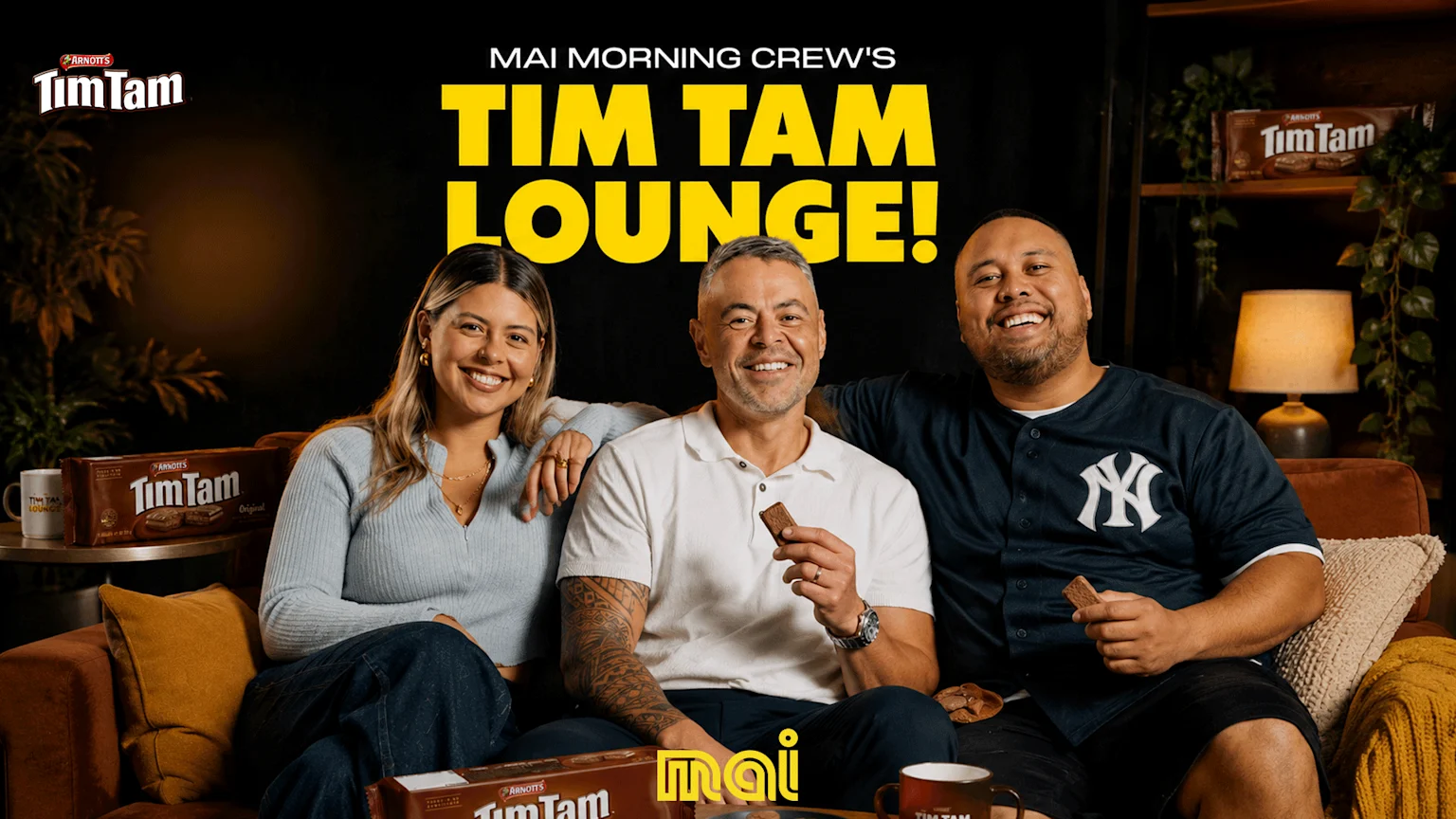 Three smiling people relax on a sofa, holding Tim Tam biscuits, surrounded by Tim Tam products, with "Mai Morning Crew's Tim Tam Lounge!" text above them.