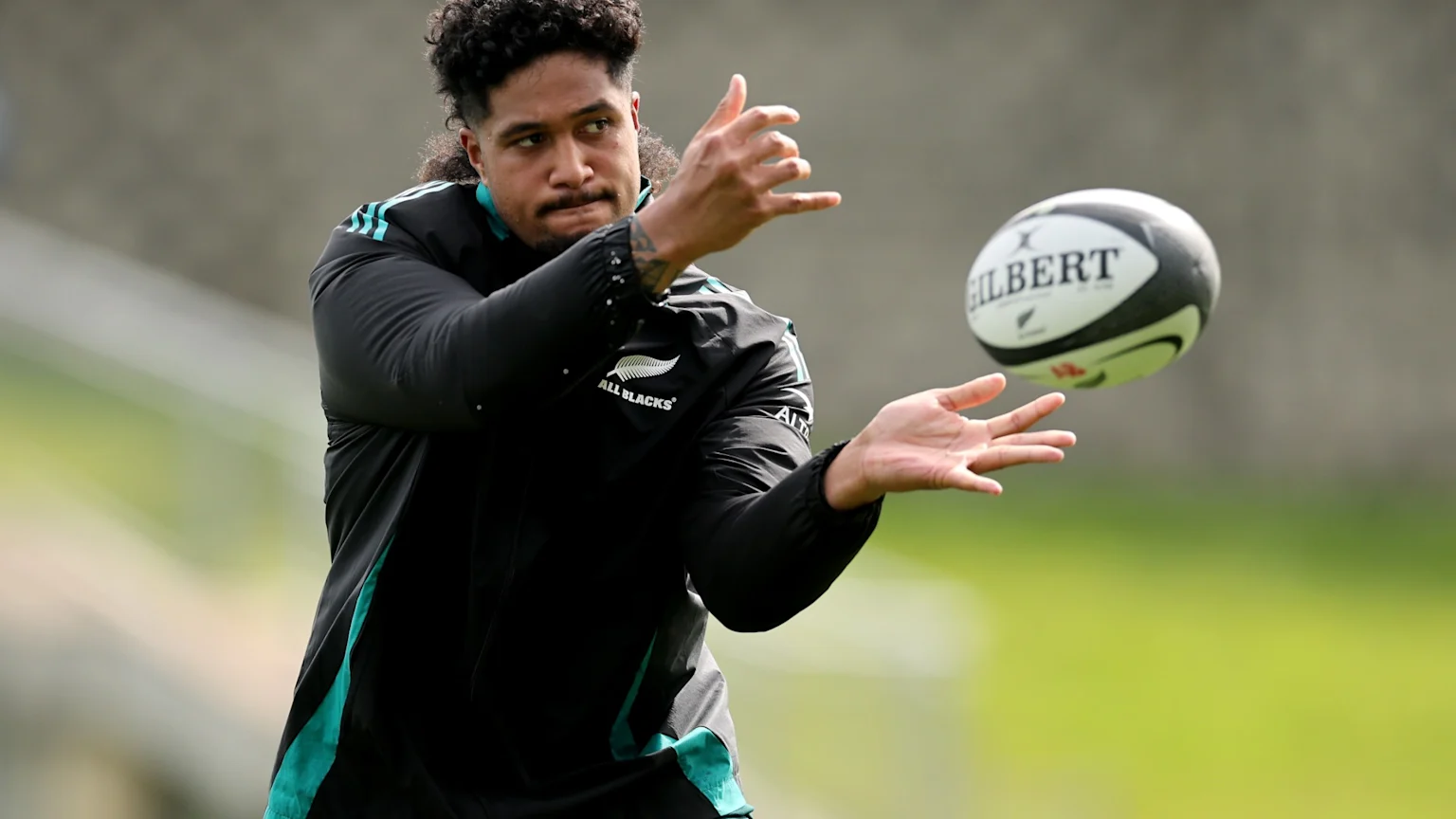 An All Blacks rugby player in a black training top with teal details looks intently at a Gilbert rugby ball in mid-air, hands open to receive it, on a green field.