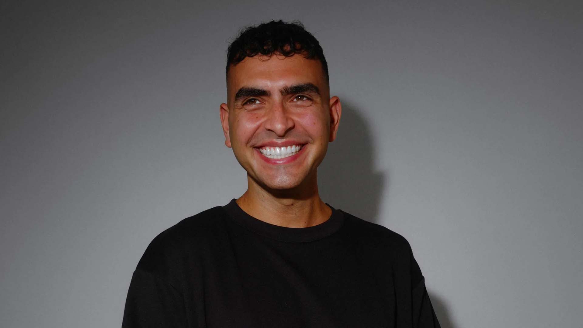 A light-skinned man with dark, curly hair and a broad smile, wearing a black t-shirt, looks upwards and to the right against a plain grey background.