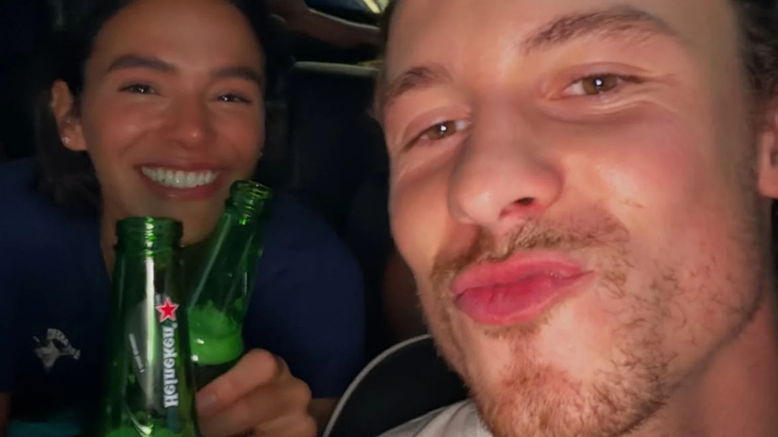 A close-up of a man with light stubble puckering his lips, with a broadly smiling woman holding two green Heineken beer bottles visible behind him.