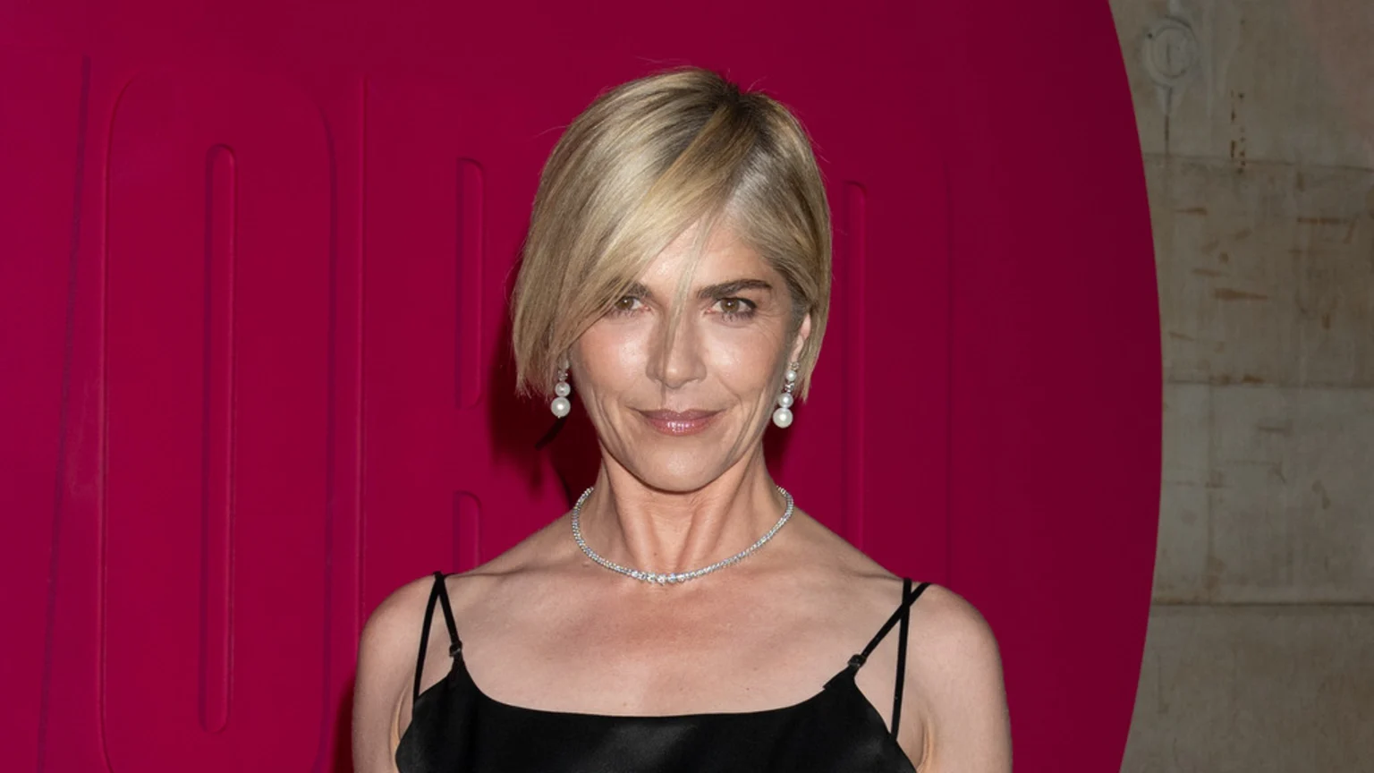 A woman with short, blonde bob hair and light brown eyes smiles gently, wearing a black slip dress, diamond necklace, and pearl drop earrings against a magenta background with embossed patterns.