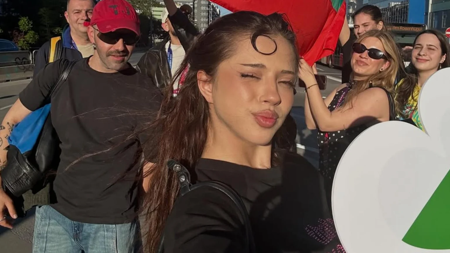A young woman takes a selfie with a kissing face, foregrounding a crowd including a man in a red cap and sunglasses, and a woman holding a red and green flag, all outdoors.