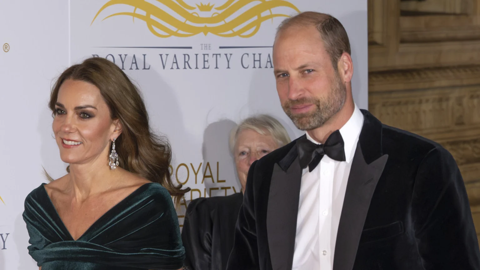 Catherine, Princess of Wales, wears a dark green velvet off-the-shoulder dress with dangling diamond earrings, smiling warmly. William, Prince of Wales, stands beside her in a black velvet tuxedo, white shirt, and black bow tie. Behind them is a white and gold banner for The Royal Variety Charity at an indoor formal gala event.
