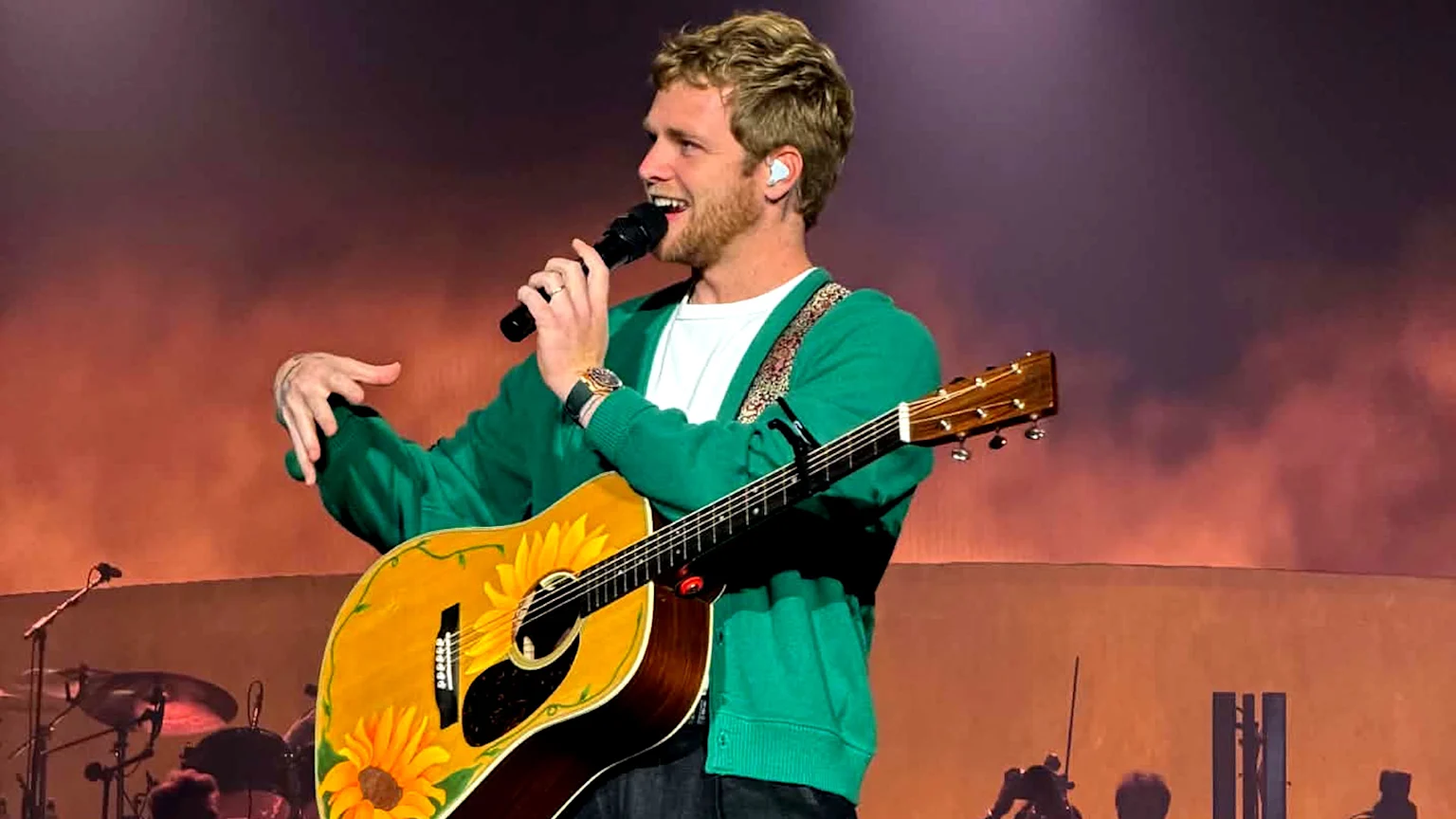 A smiling man with blonde hair and a beard, wearing a green cardigan, holding a microphone, and playing a sunflower-decorated acoustic guitar on stage.