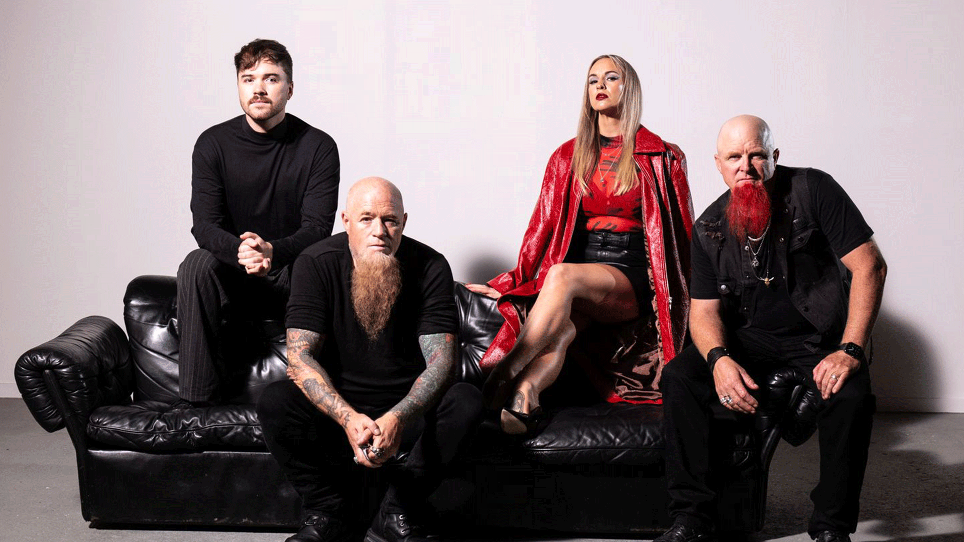 Four band members, including a woman in a red coat and a man with a bright red beard, pose on and around a black leather couch against a white background.
