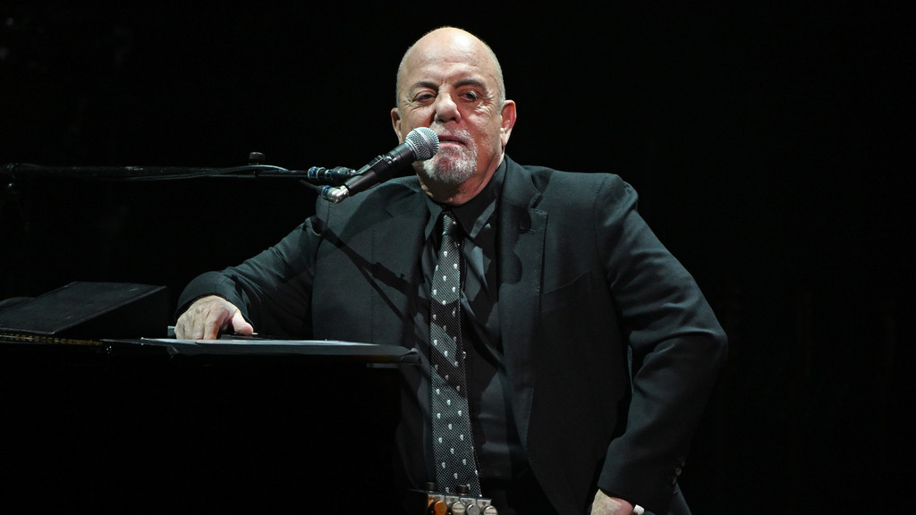 Billy Joel, bald with a goatee, sits at a piano in a black suit and polka-dotted tie, looking at the camera with a microphone in front of him.