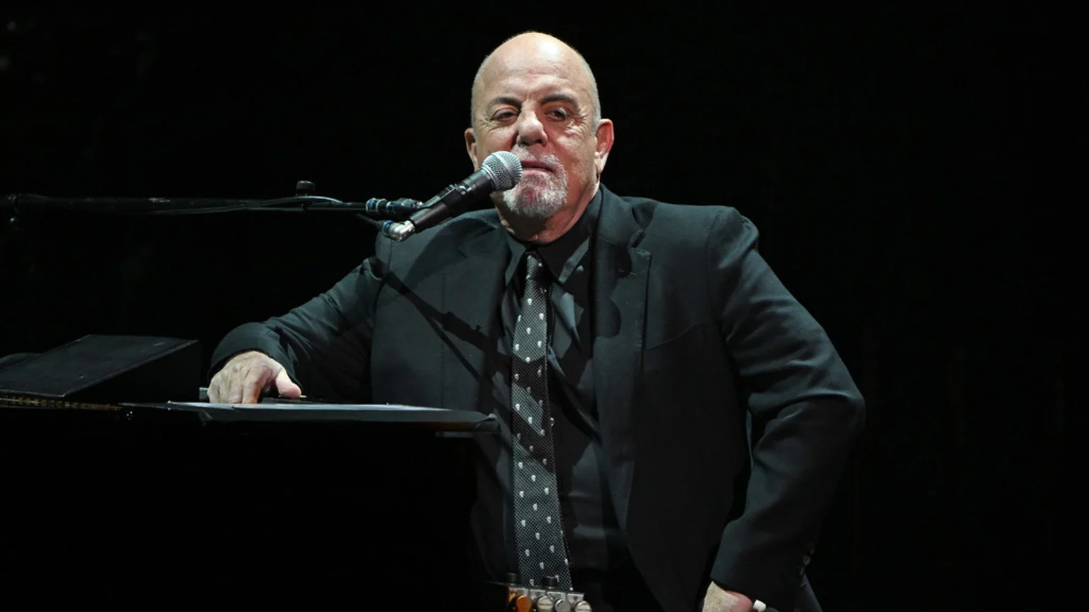 Billy Joel, bald with a goatee, sits at a piano in a black suit and polka-dotted tie, looking at the camera with a microphone in front of him.
