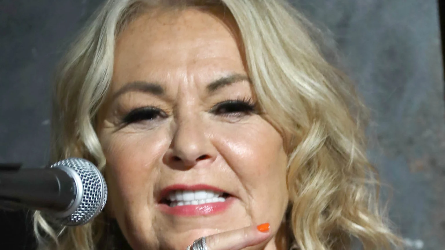 A blonde woman with makeup and visible wrinkles smiles, showing her teeth, with a microphone near her face and an orange-painted finger touching her chin.
