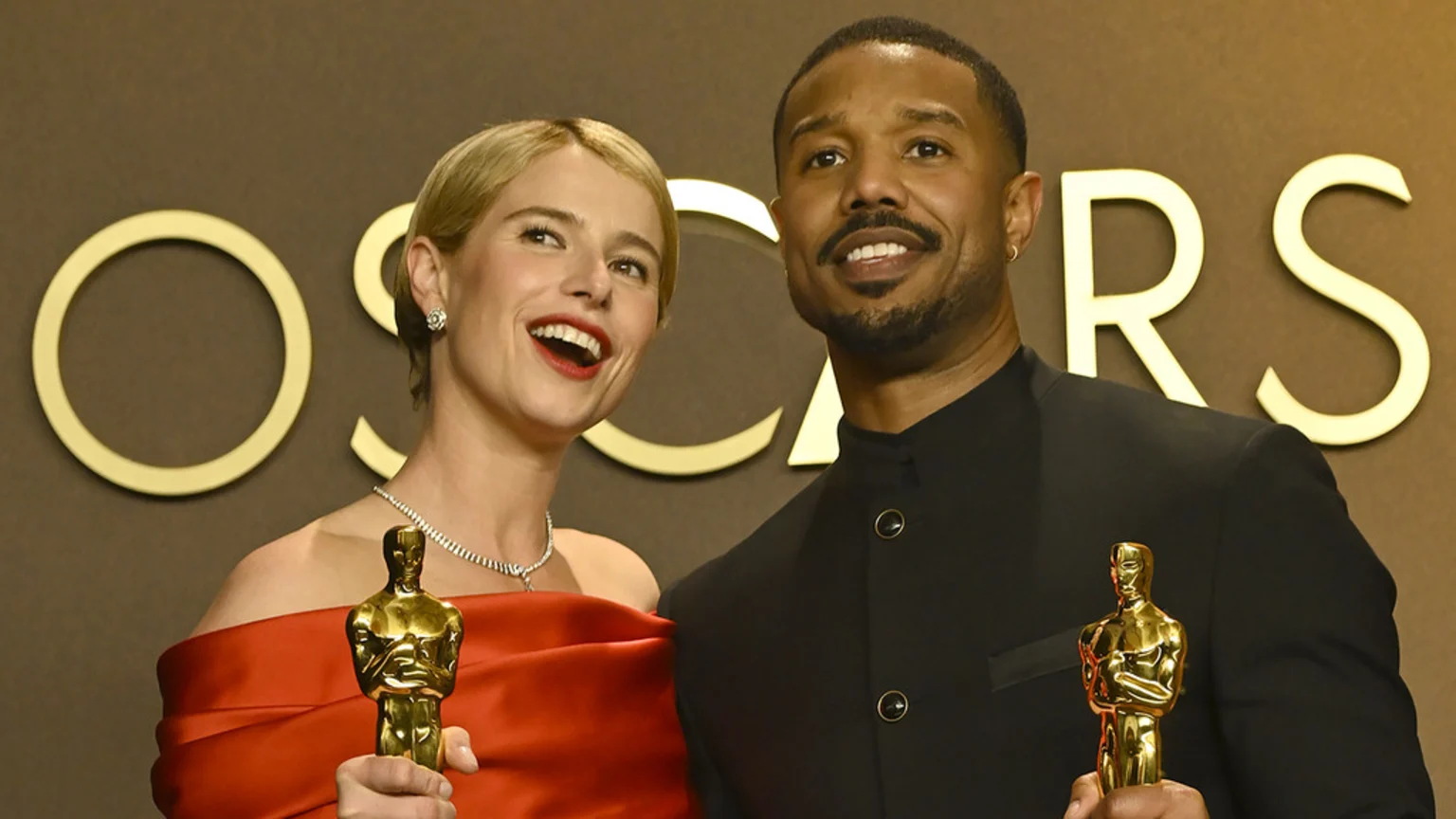 A woman in a red dress and a man in a black suit smile while holding golden Oscar statues in front of an "OSCARS" backdrop.