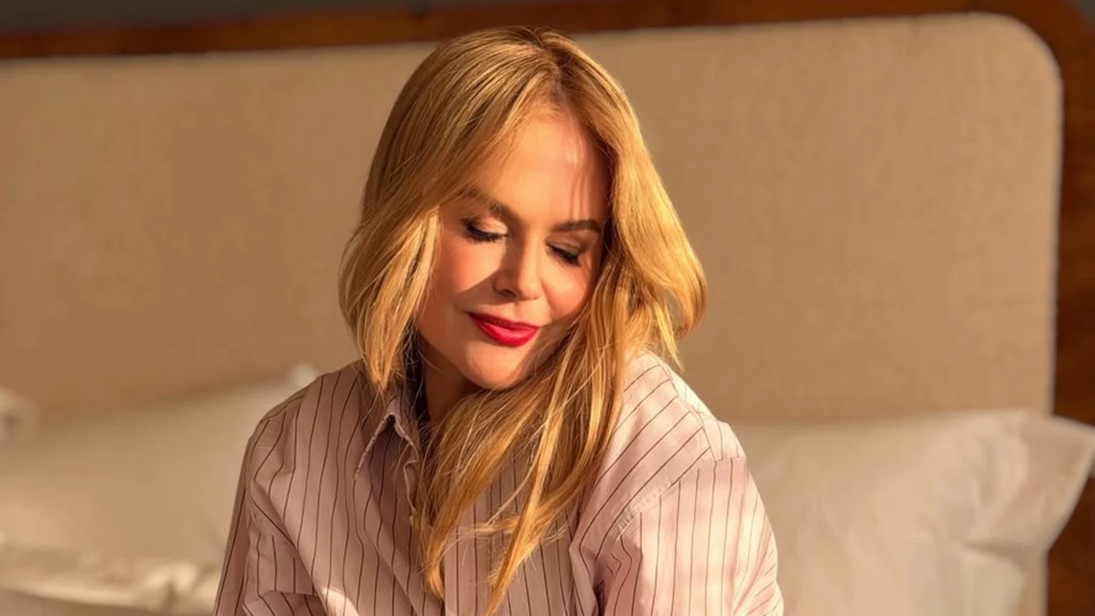 Nicole Kidman with blonde hair and red lipstick, smiling serenely with closed eyes, wearing a light-colored striped shirt. She is bathed in warm, golden light against a soft, blurry background.