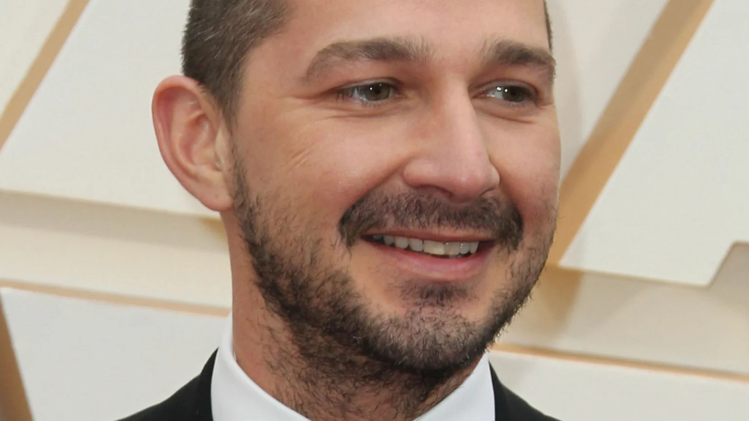 Close-up of actor Shia LaBeouf smiling, with short hair and a beard, looking slightly to the right, wearing a suit jacket and white shirt.