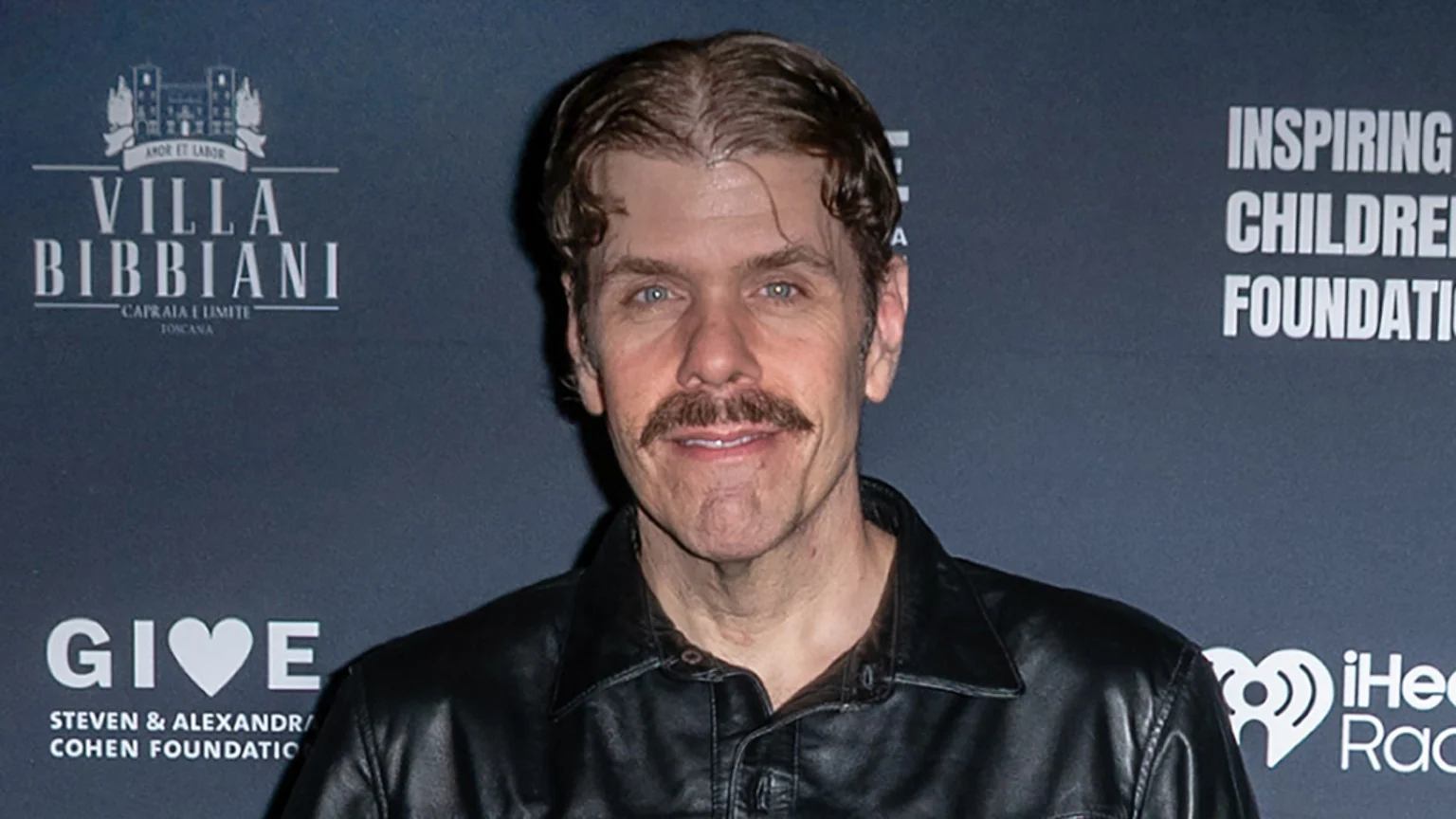 Headshot of a man with a mustache, wavy brown hair, and blue eyes, wearing a black leather jacket, posing in front of a dark background with various logos.