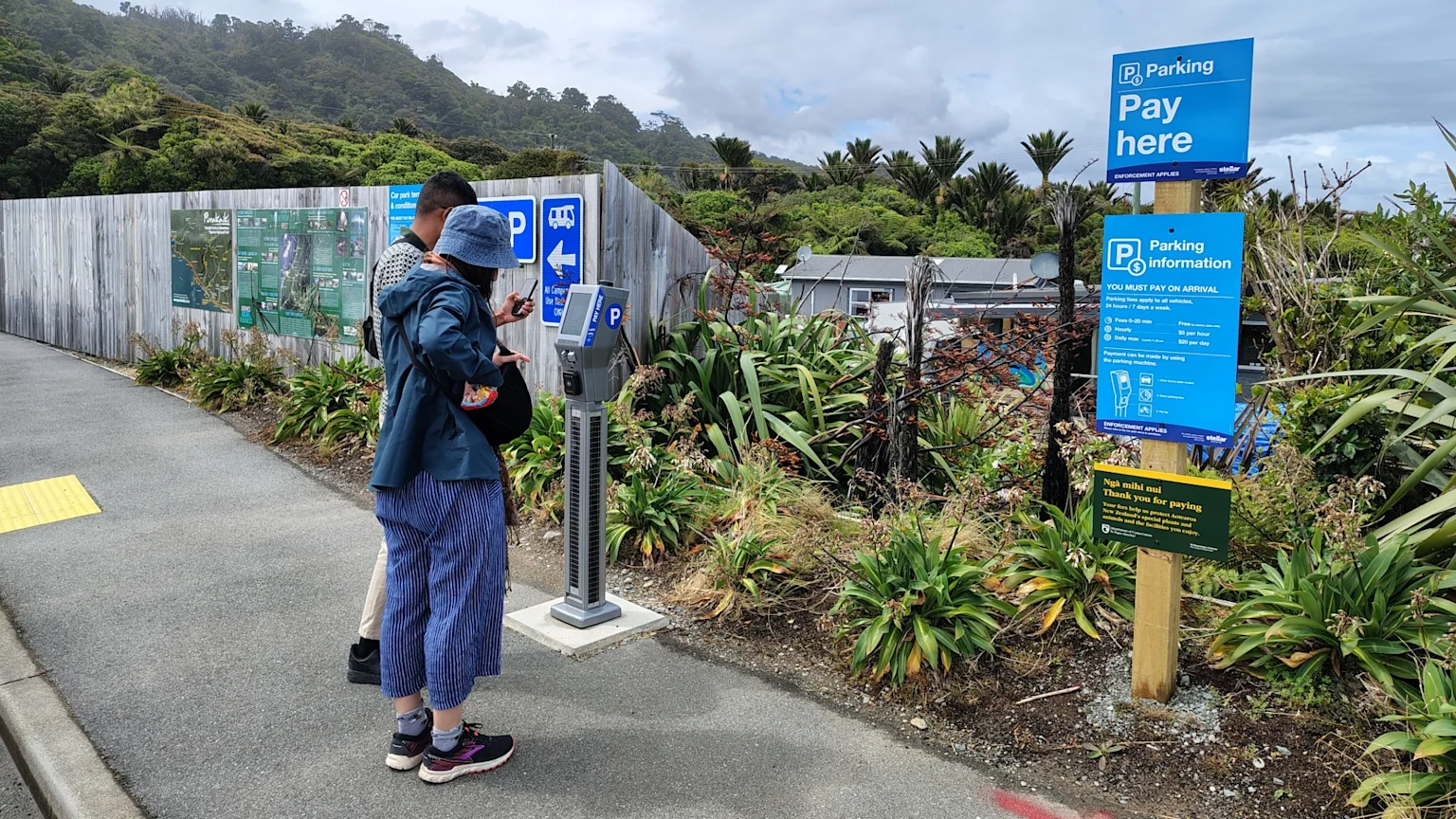 Two people stand at a parking payment machine next to a wooden fence with maps and parking signs, surrounded by lush green vegetation and hills under a cloudy sky.