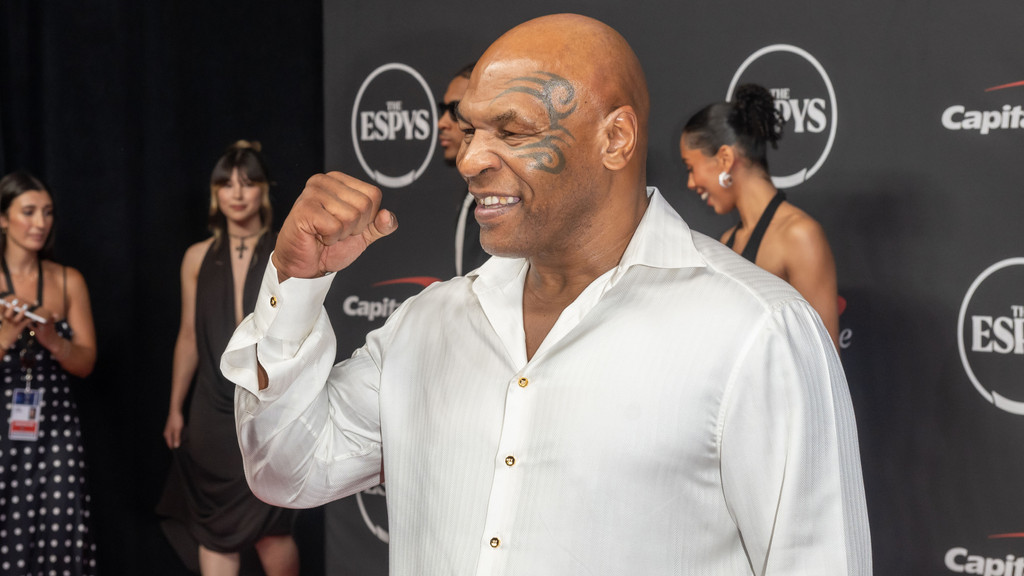 Mike Tyson, smiling with his face tattoo, stands on a red carpet at the ESPYs, wearing a white shirt with gold buttons and clenching his right fist, with blurred people and logos in the background.
