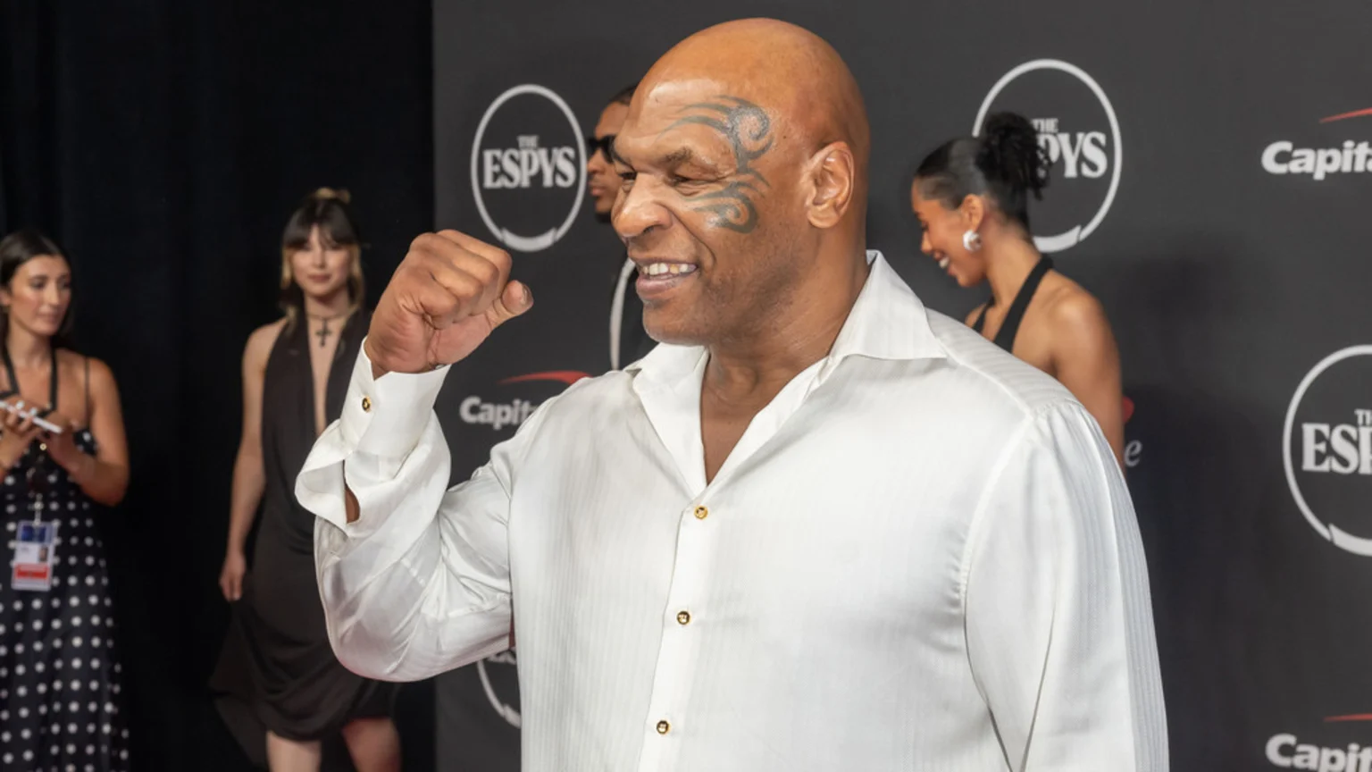 Mike Tyson, smiling with his face tattoo, stands on a red carpet at the ESPYs, wearing a white shirt with gold buttons and clenching his right fist, with blurred people and logos in the background.