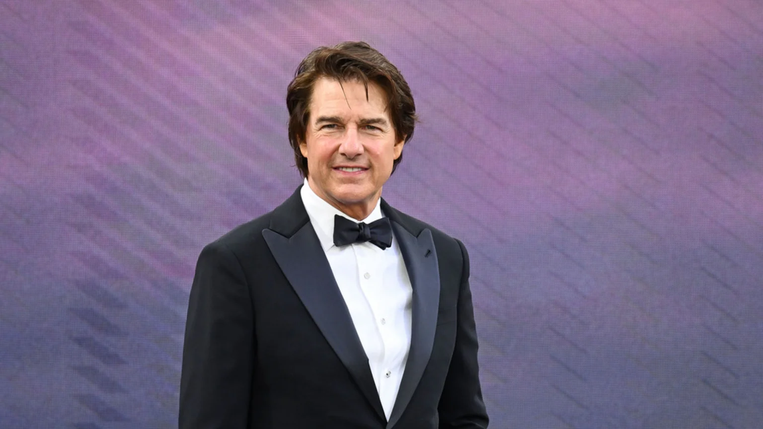 Tom Cruise smiles at the camera, wearing a black tuxedo with a bow tie and white shirt, against a blurred purple background.