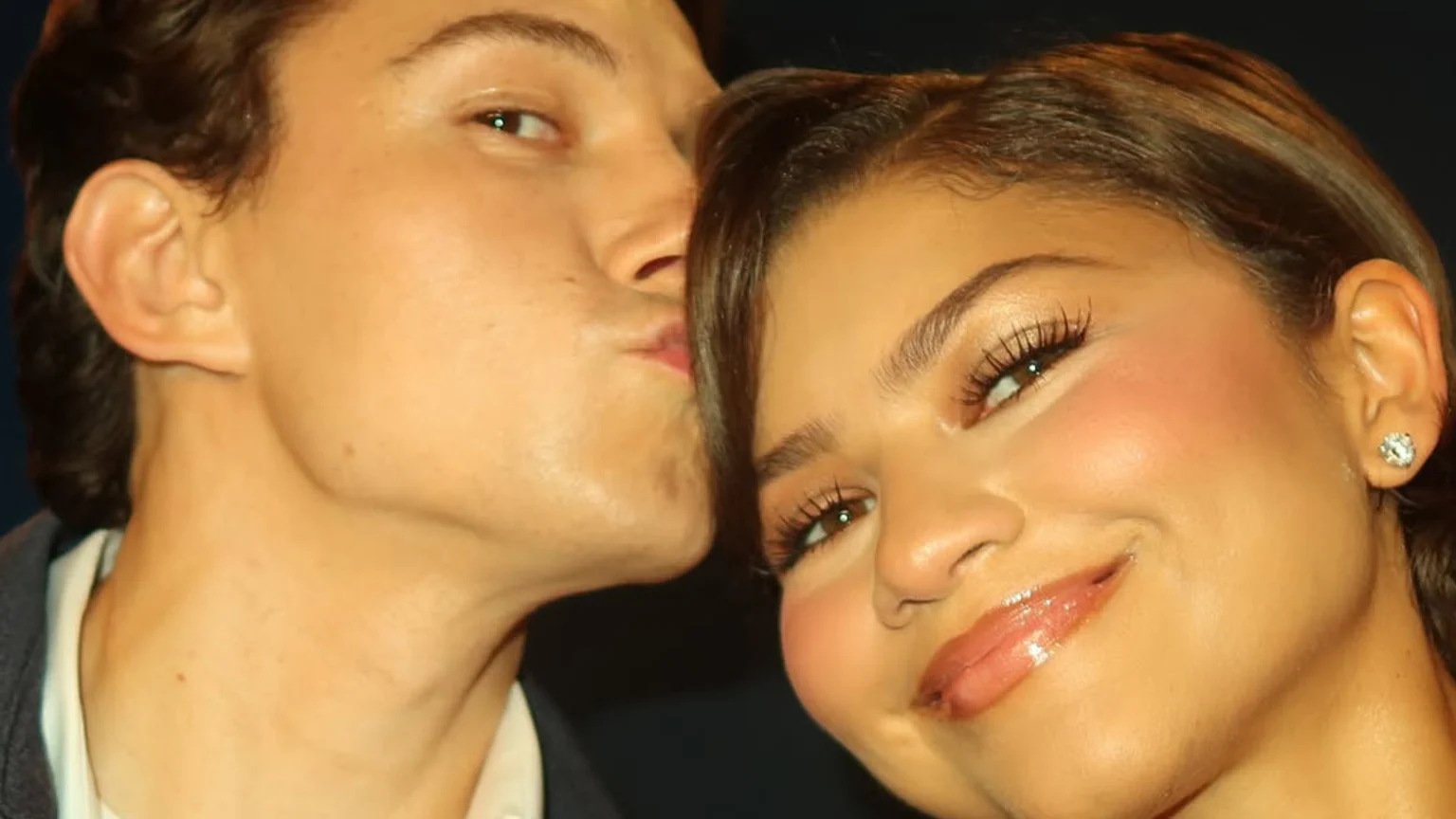 Close-up portrait of a man with puckered lips leaning towards a smiling woman with glossy lips and a stud earring, who looks at the camera.
