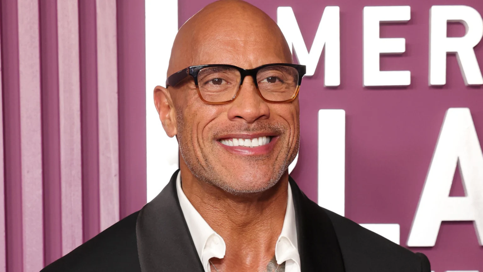 Dwayne Johnson, bald and wearing glasses, smiles broadly in a dark suit, against a purple background with white vertical stripes and white text.