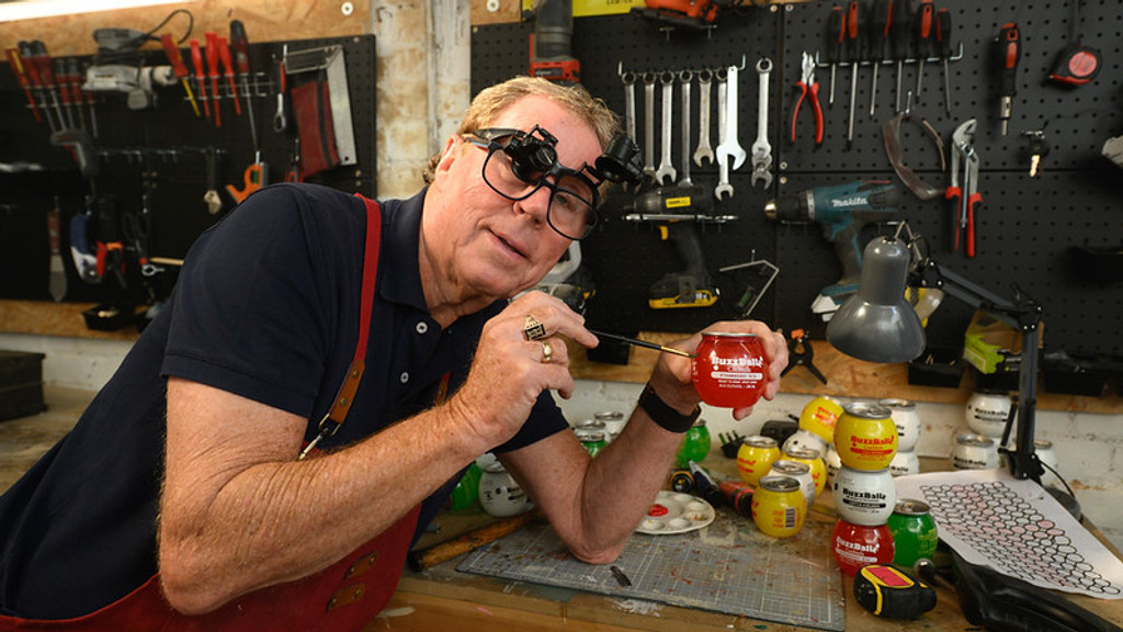A man wearing magnifying glasses and an apron paints a red "Buzzballz" can on a workbench, surrounded by other colorful cans, paints, and tools hanging on a pegboard wall.