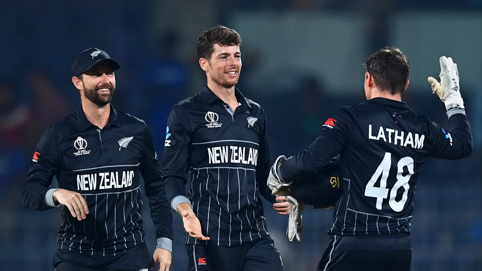 Three New Zealand cricket players in black uniforms, two smiling, interact as one player's back reveals "LATHAM" and "48."