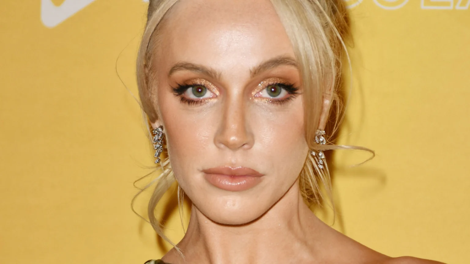 A close-up of Alex Cooper with blonde hair, green eyes, shimmery golden eyeshadow, and nude lips, wearing diamond chandelier earrings against a yellow background.