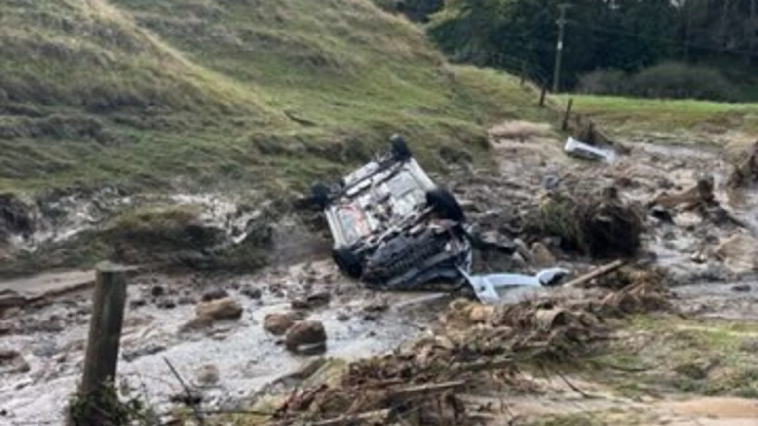 An overturned dark-colored vehicle lies partially submerged in a muddy gully filled with debris, with a grassy hill in the background.