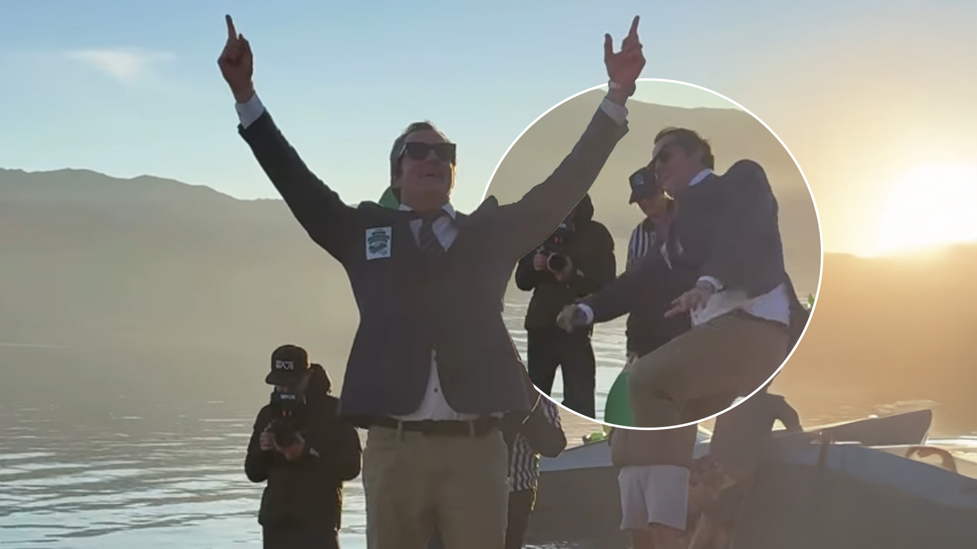 A man in a suit, tie, sunglasses, and khaki pants stands triumphantly with arms raised on a calm lake at sunset, mountains in the background; a circular inset shows him mid-action on a boat.