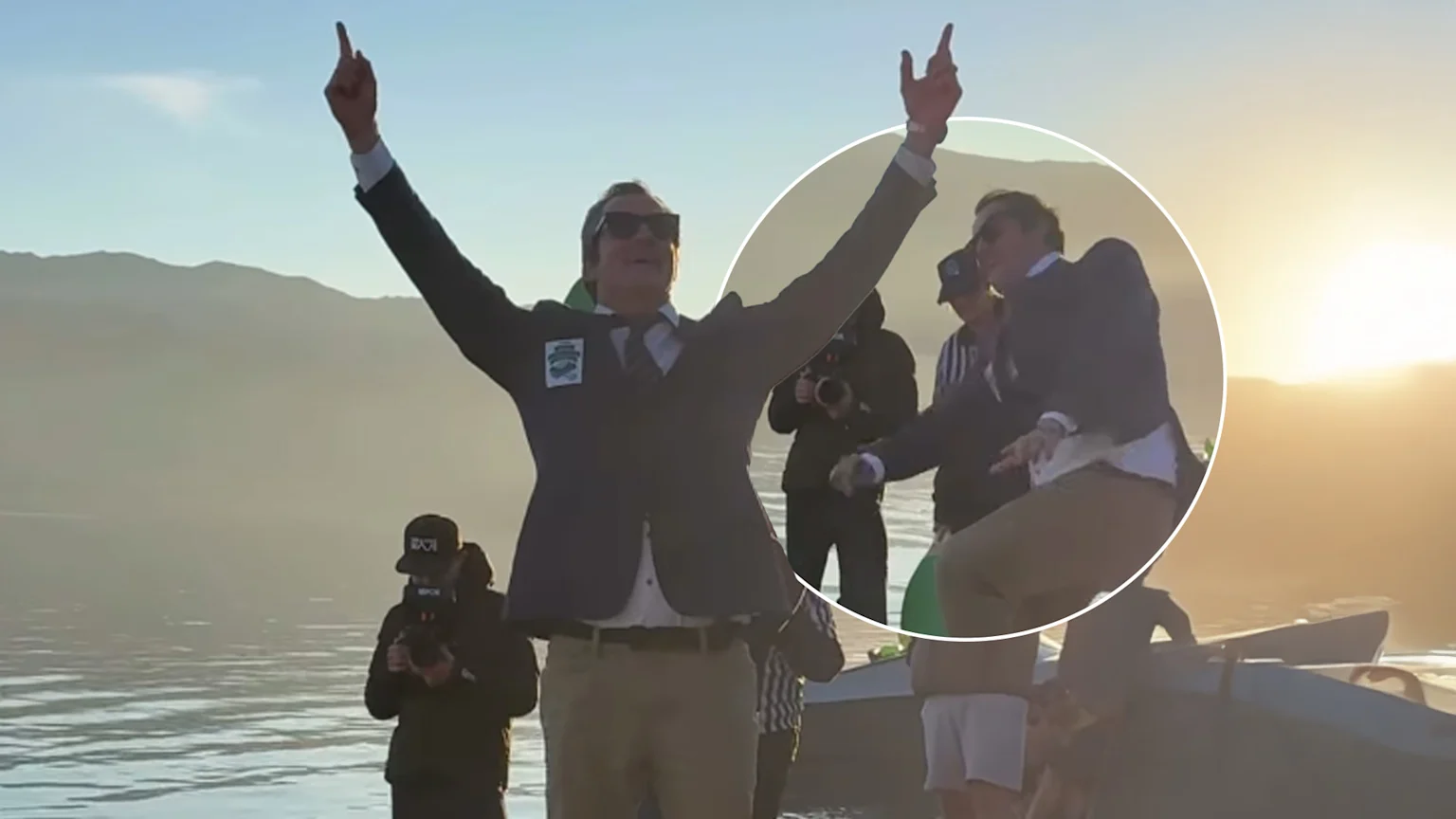 A man in a suit, tie, sunglasses, and khaki pants stands triumphantly with arms raised on a calm lake at sunset, mountains in the background; a circular inset shows him mid-action on a boat.