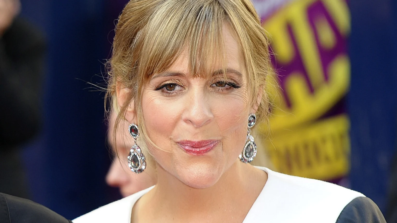 Close-up portrait of a middle-aged blonde woman wearing white and black attire with teardrop-shaped dangling earrings. She smiles slightly, set against a colorful, blurred event backdrop with abstract shapes.
