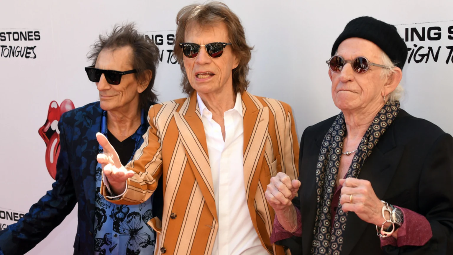The Rolling Stones members Ronnie Wood, Mick Jagger, and Keith Richards stand side-by-side outdoors at an event with the band's logo in the background.