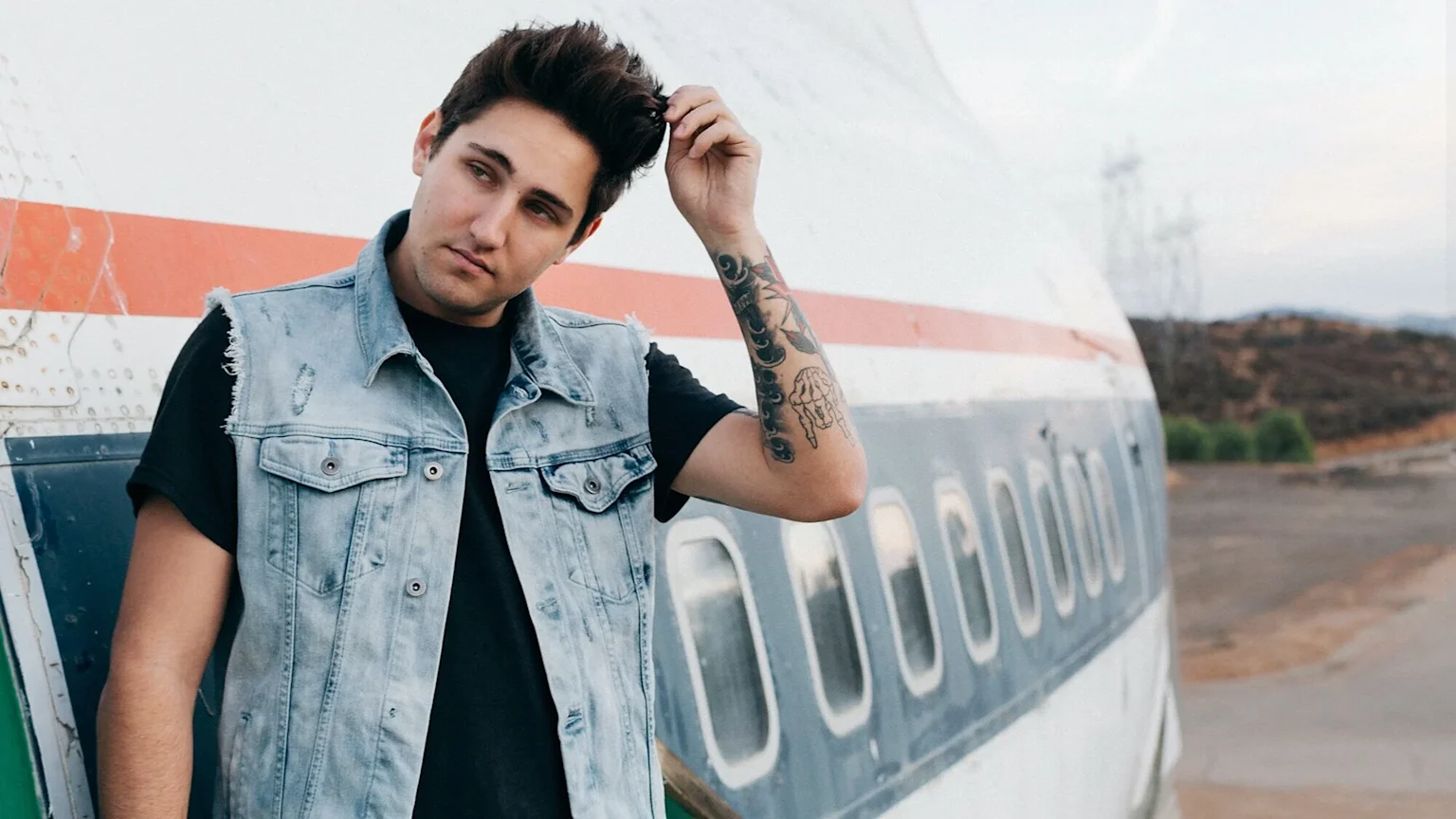A young man with dark hair and tattoos on his left arm, wearing a light blue distressed denim vest and black t-shirt, stands next to an old airplane, touching his hair.