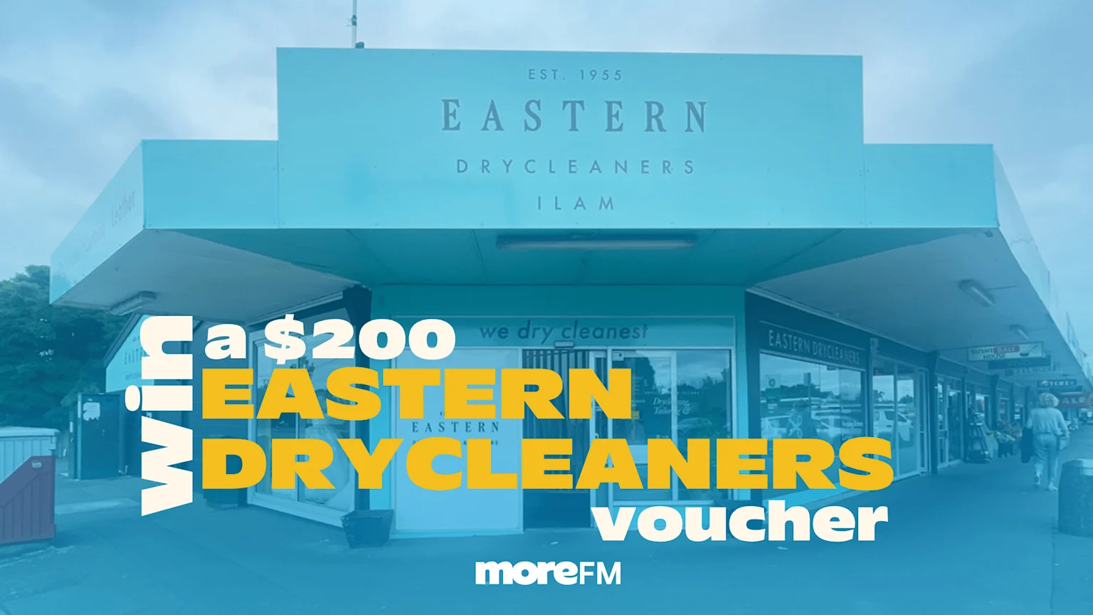 A blue-green building housing "Eastern Drycleaners" with a prominent text overlay advertising "Win a $200 Eastern Drycleaners voucher moreFM" on an overcast day.