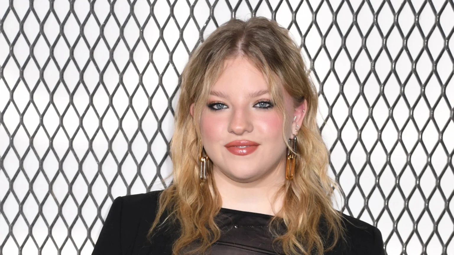Francesca Scorsese, a young woman with long blonde hair and light makeup, looks at the camera wearing a dark top and large golden earrings against a black diamond-patterned grid.