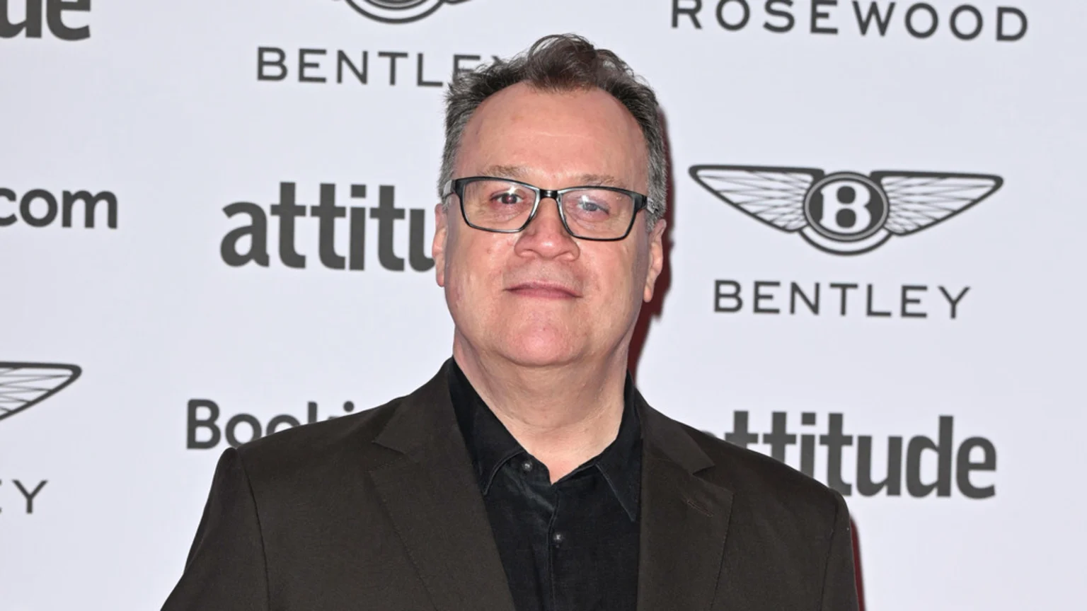 A man with glasses and a dark suit stands in front of a white background with "attitude" and Bentley logos.