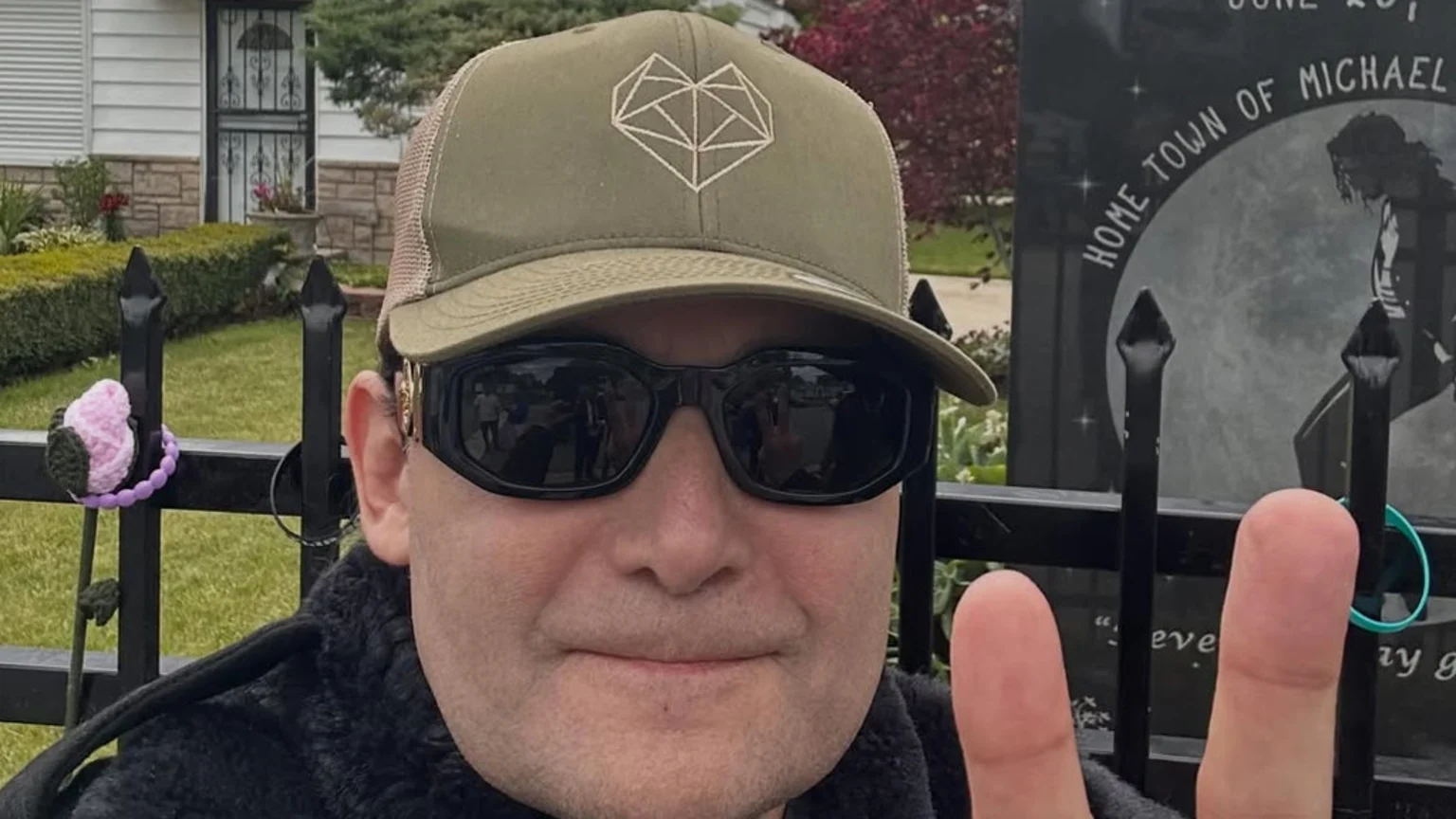 A man in a green cap and black sunglasses smiles and holds up two fingers in a peace sign, with a black fence and a Michael Jackson 'Home Town' monument in the background.