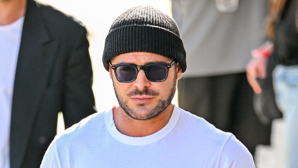 Zac Efron with a beard, wearing a black beanie, dark sunglasses with orange accents, and a white t-shirt, looking forward.
