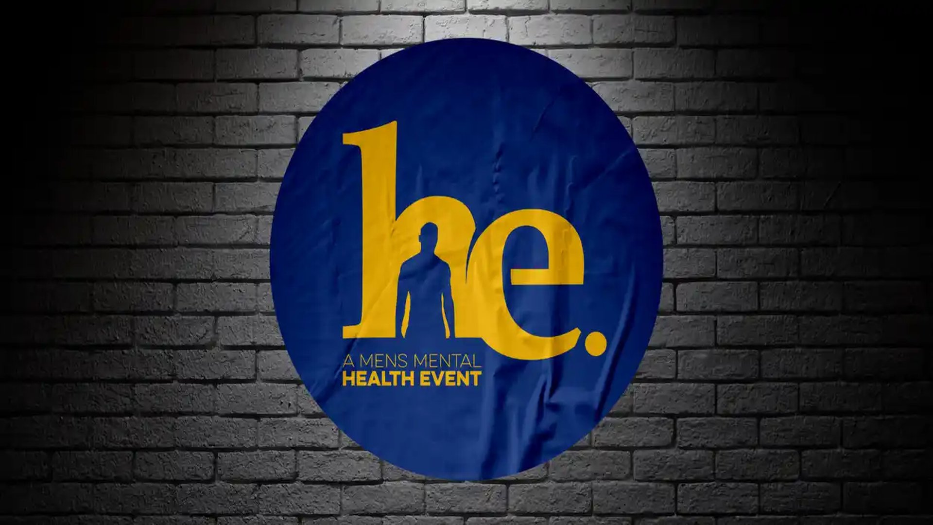 A circular blue banner with the word "he." in large yellow letters, where the "h" incorporates a human silhouette, and "A MENS MENTAL HEALTH EVENT" below, against a dark brick wall.