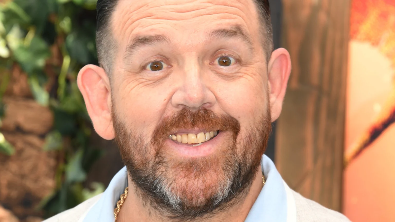 Nick Frost smiles at the camera, showing a reddish-brown beard, short dark hair, and a gold chain over a light shirt, with a blurred natural background.