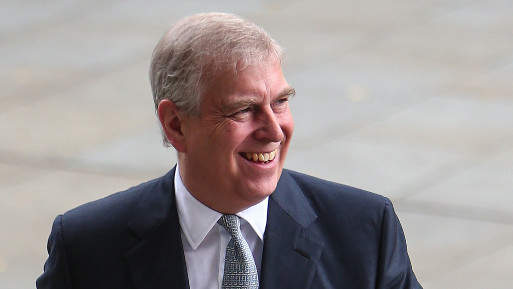 A man with short grey hair, wearing a dark suit and patterned tie, smiles broadly while looking towards the right side of the frame.