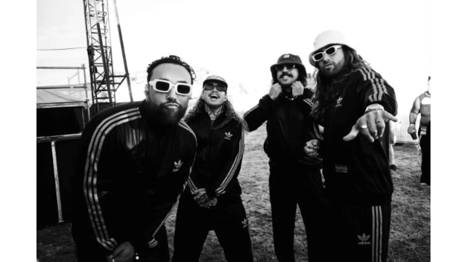 A black and white photo features four men in black Adidas tracksuits and sunglasses, posing outdoors; one man gestures towards the viewer.