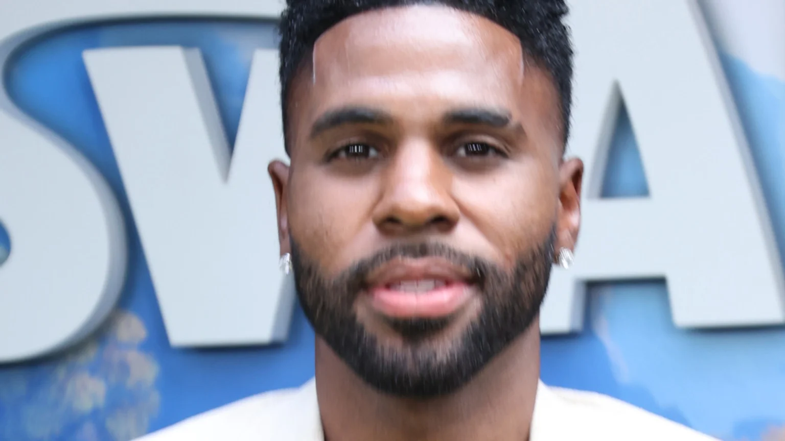 A close-up headshot of Jason Derulo, a man with dark skin and a neat beard, smiling faintly at the camera. Large, blurry white letters spelling "SWAN" are visible on a blue background behind him.