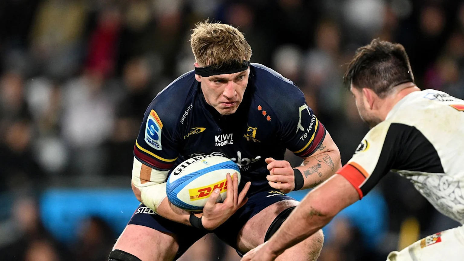 A male rugby player wearing a dark blue Highlanders jersey with multiple sponsor logos holds a Gilbert rugby ball, fends off an opponent in a white and black jersey, and is taped on the forearms. The game takes place at night in an outdoor stadium with artificial lighting.