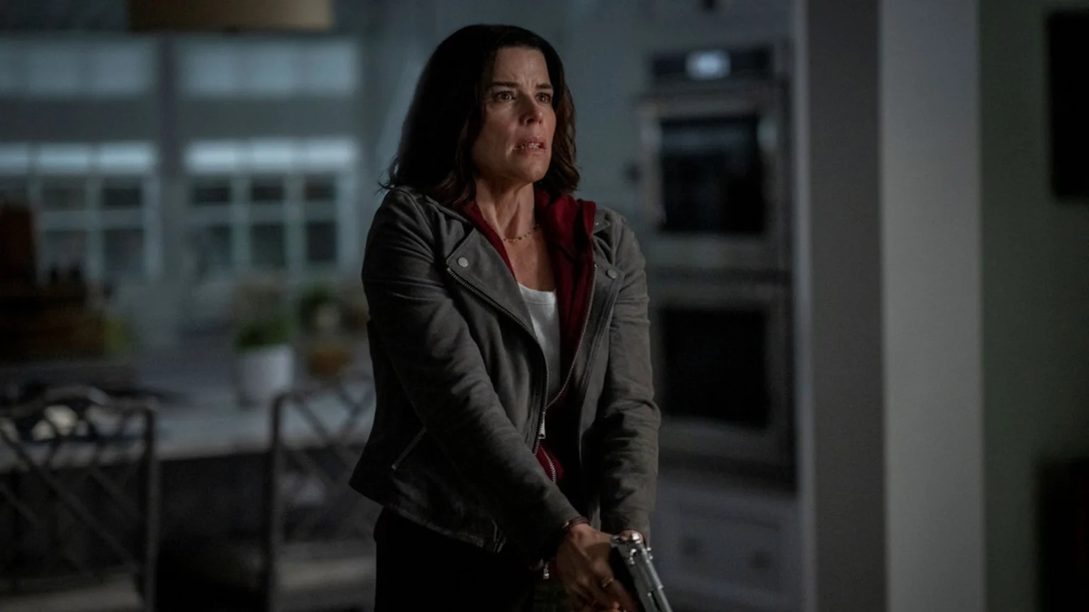Neve Campbell, looking concerned, holds a silver handgun with both hands, wearing a gray jacket over a red hoodie in a dimly lit indoor setting.