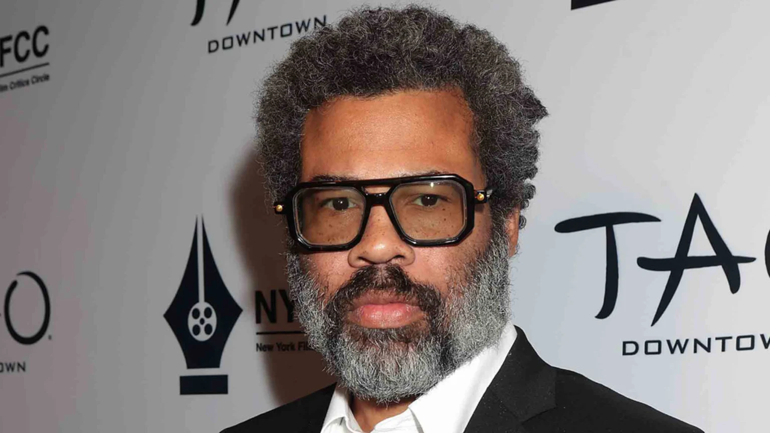 Jordan Peele, with graying curly hair, a gray beard, and distinctive large black glasses, looks directly at the camera, wearing a white shirt and black suit jacket.