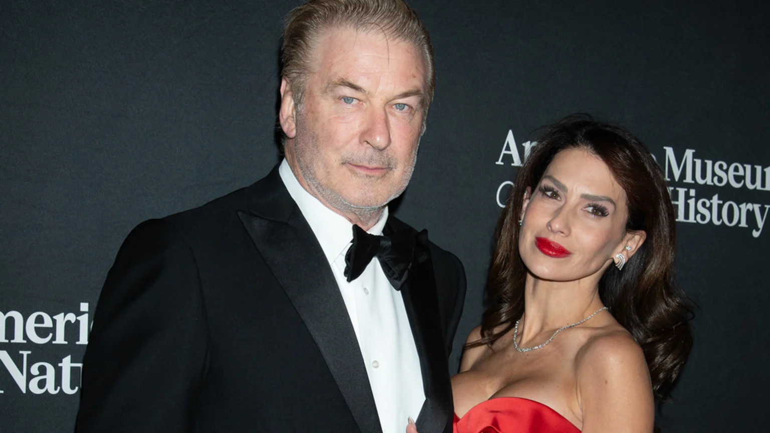 Alec Baldwin in a tuxedo and Hilaria Baldwin in a red strapless gown and diamond necklace pose against a dark background.