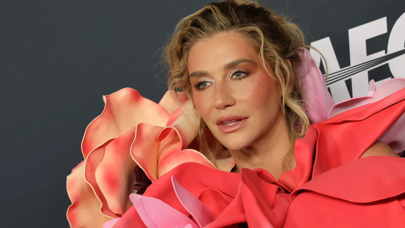 Close-up of singer Kesha with blonde, curly hair, wearing an elaborate, sculptural dress made of large pink and red fabric petals, against a dark background with a partial white logo.