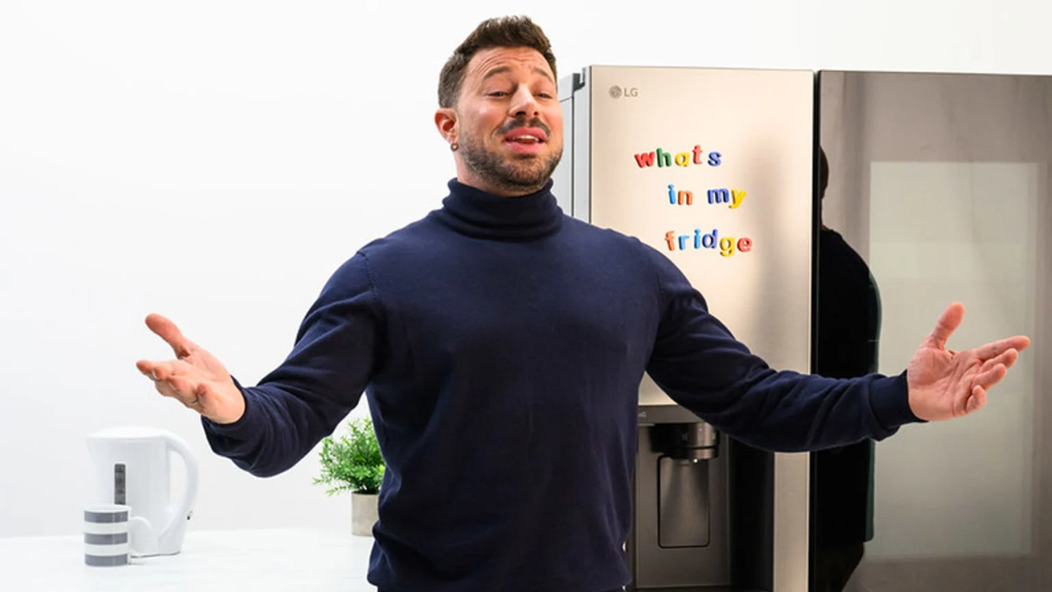A man in a dark blue turtleneck sweater stands in front of an LG refrigerator, gesturing with open hands, with magnetic letters on the fridge spelling "whats in my fridge".